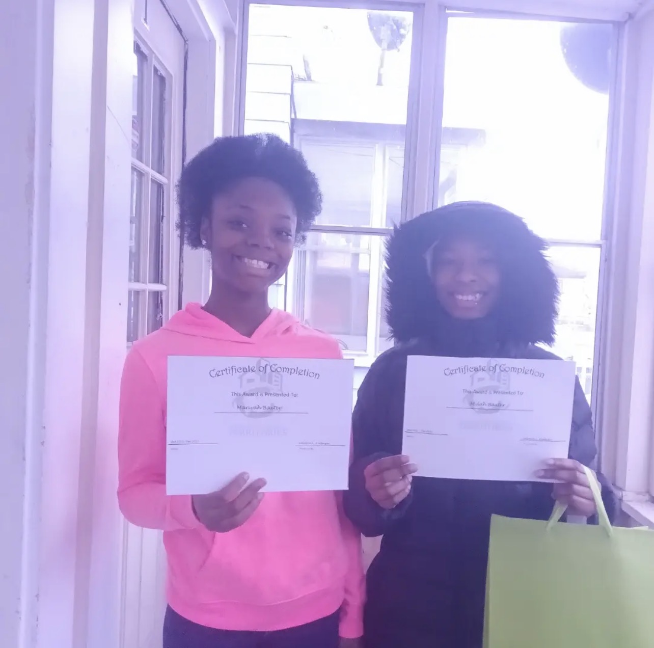 Two smiling graduates with their certificates by a bright window